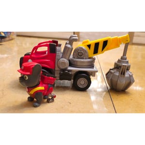 May include: A red and gray toy tow truck with a yellow crane arm and a gray wrecking ball. A small gray and red cartoon dog figure in a firefighter uniform stands next to the truck. The toy is on a light brown surface.