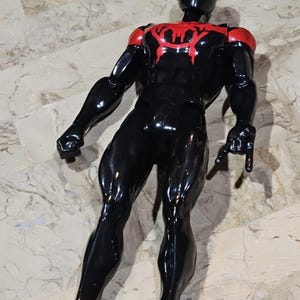 May include: A black and red action figure of a superhero. The figure has a black body with red accents on the shoulders and chest. The superhero's mask is black with white eyes. The figure is posed with its arms outstretched.
