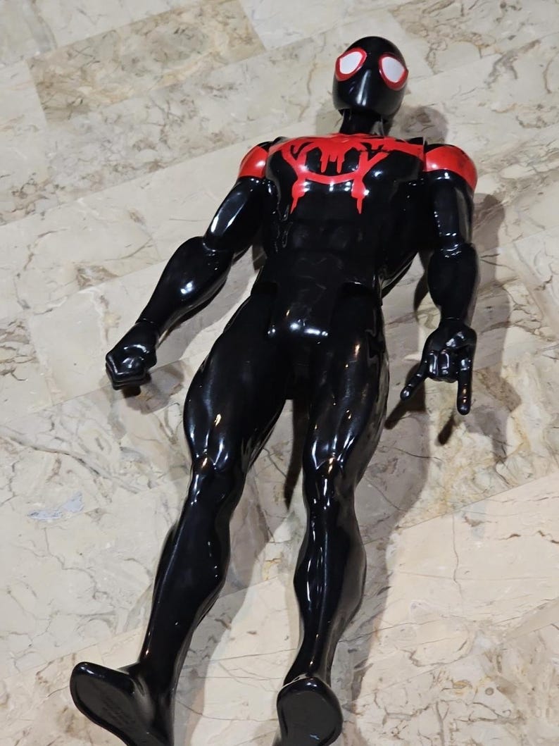 May include: A black and red action figure of a superhero. The figure has a black body with red accents on the shoulders and chest. The superhero's mask is black with white eyes. The figure is posed with its arms outstretched.