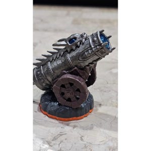 Puede incluir: Un cañón de juguete plateado con diseño de cabeza de dragón, detalles azules con forma de gema y una base de rueda marrón. El cañón se asienta sobre una base negra y naranja. El juguete es probablemente un objeto de colección o parte de un juego más grande.