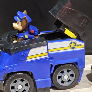 May include: Blue and black toy truck with a yellow stripe and a Paw Patrol logo. A brown and black toy dog figure is sitting in the driver's seat. The truck has a removable top and a back door that opens.