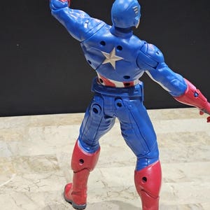 May include: A blue and red action figure of Captain America with a star on his chest. The figure is posed with one arm raised and the other arm extended to the side. The figure is standing on a tiled surface.
