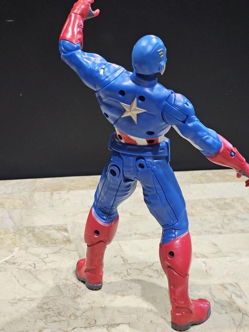 May include: A blue and red action figure of Captain America with a star on his chest. The figure is posed with one arm raised and the other arm extended to the side. The figure is standing on a tiled surface.