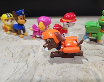 May include: A set of six colorful plastic toy figurines of characters from the children's television show Paw Patrol. The figurines are all wearing different outfits and accessories, including a police officer, a firefighter, a construction worker, a pilot, a farmer, and a veterinarian.