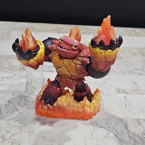 Puede incluir: Una figura de juguete elemental de fuego roja y naranja con una base negra y llamas. La figura está de pie sobre una base de llamas naranjas.