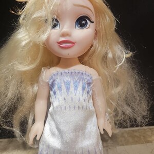 May include: A blonde doll wearing a white and blue dress with a snowflake pattern. The doll has blue eyes and a smiling face.