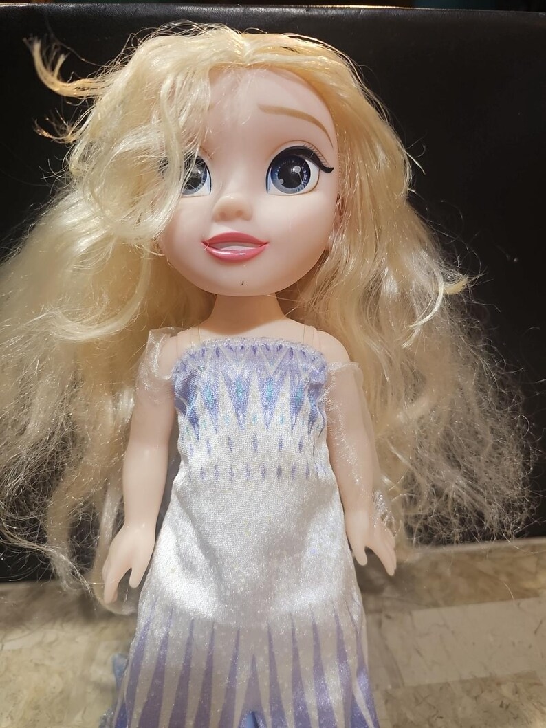 May include: A blonde doll wearing a white and blue dress with a snowflake pattern. The doll has blue eyes and a smiling face.