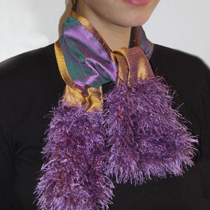 Op de afbeelding: Een paarse en gouden sjaal met een glanzend uiterlijk. De sjaal heeft een combinatie van paarse en gouden stoffen, met een pluizige, gefranjerde textuur aan de uiteinden. De tekst "PURPLE & GOLD SCARF" staat onderaan.