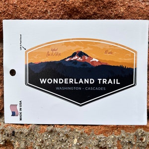 May include: A sticker featuring a stylized illustration of a mountain range with a peak in the background. The sticker is orange, yellow, and blue with the text "WONDERLAND TRAIL WASHINGTON - CASCADES" in black. The sticker is made in the USA.