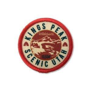 Könnte beinhalten: Ein runder roter und weißer Aufnäher mit dem Text "Kings Peak Scenic Utah" und einer Bergkette als Illustration im Inneren. Der Text "Wilderness" befindet sich über der Bergkette und die Zahl "13334" unter der Bergkette.