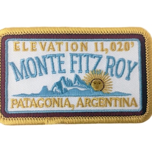 May include: A white embroidered patch with a gold border and a maroon inner border. The patch features the text "ELEVATION 11,020' MONTE FITZ ROY PATAGONIA, ARGENTINA" and a stylized image of a mountain range with a sun.
