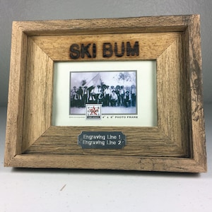 May include: A rustic wooden photo frame with the word "SKI BUM" carved at the top. The frame holds a black and white photo and a small metal plate for engraving. The frame is designed for a 4" x 6" photo.