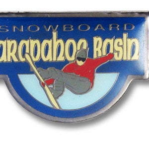 May include: A blue and silver pin with the words "SNOWBOARD" and "arapahoe basin" in yellow lettering. The pin features a skier in a red jacket and gray pants. The skier is on a snowboard.