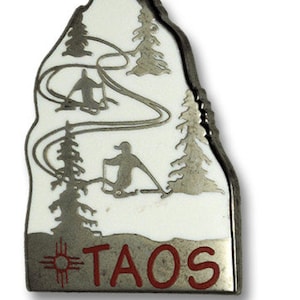 Op de afbeelding: Een zilveren metalen pin met een witte emaille achtergrond waarop een berglandschap met skiërs en het woord "TAOS" in rood is afgebeeld.