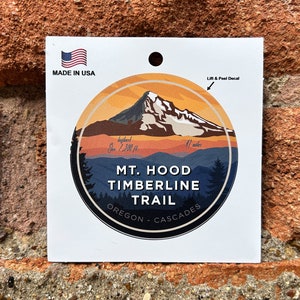 Pode incluir: Um adesivo redondo com uma ilustração colorida do Monte Hood, Oregon. O adesivo apresenta um céu de pôr do sol com um pico de montanha coberto de neve. O texto "MT. HOOD TIMBERLINE TRAIL OREGON - CASCADES" está impresso no adesivo.