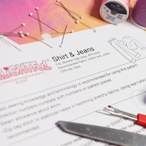 May include: A white sheet of paper with the text "Shirt & Jeans" and a line drawing of a shirt and jeans. The text "Dlightful" is in a colorful font at the top of the page. The paper is surrounded by sewing supplies, including pins, thread, and a seam ripper.