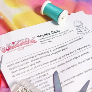 May include: A white sheet of paper with the text "Dollightful Hooded Cape" printed on it. The text is in a black font with a red heart and a white snowflake. The paper is lying on a pink and yellow fabric with a spool of turquoise thread.