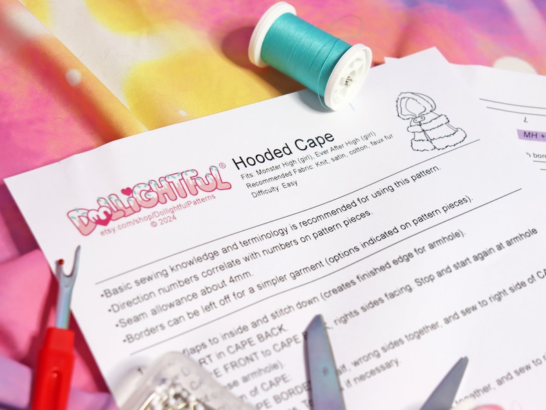 May include: A white sheet of paper with the text "Dollightful Hooded Cape" printed on it. The text is in a black font with a red heart and a white snowflake. The paper is lying on a pink and yellow fabric with a spool of turquoise thread.