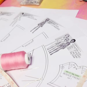 May include: A sewing project with a pink spool of thread, a measuring tape, and a pattern for a bodice. The pattern includes instructions for a Monster High doll, with the text "Monster High Gen 3 Standard" and "Monster High Gen 1 Standard".