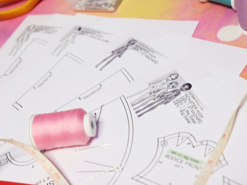 May include: A sewing project with a pink spool of thread, a measuring tape, and a pattern for a bodice. The pattern includes instructions for a Monster High doll, with the text "Monster High Gen 3 Standard" and "Monster High Gen 1 Standard".