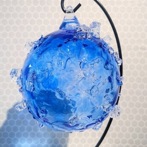 May include: A round, blue glass ornament with a textured surface and clear, crystalline accents. The ornament is suspended from a black metal stand, creating a decorative display. The glass has a marbled appearance.