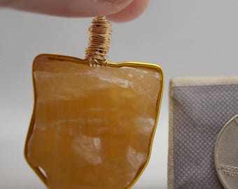 May include: A pendant labeled "A" featuring a golden-yellow stone with a gold-colored wire wrap. The pendant is held up by a person's finger. A quarter is next to the pendant for size reference.
