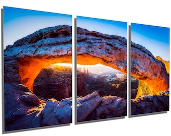 May include: A panoramic view of a natural sandstone arch in a desert landscape. The arch is illuminated by the rising sun, casting a warm orange glow on the rock formations. The sky is a vibrant blue, and the distant landscape is a hazy brown.