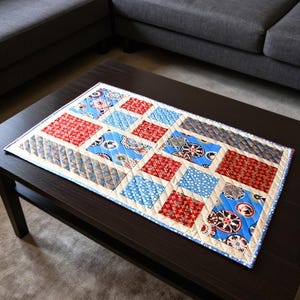 May include: A rectangular quilted table runner featuring a patchwork design of red, blue, and cream fabric squares. The pattern includes geometric shapes, floral motifs, and circular designs, all stitched together with a diagonal grid quilting pattern.