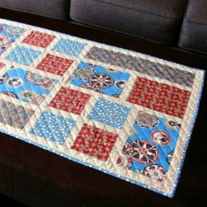 May include: A patchwork table runner with a quilted design. It features squares of red, blue, and patterned fabrics, framed by a cream-colored fabric. The text "TABLE RUNNER" is visible at the bottom.