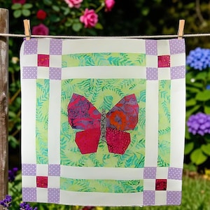 Op de afbeelding: Een vierkant stoffen quiltblok met een centrale vlinderapplicatie in tinten rood, oranje en paars. De vlinder is geplaatst tegen een lichtgroene achtergrond met een varenpatroon, omlijst door witte randen en hoekvierkanten met lavendelkleurige stippen.