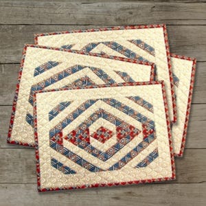 Könnte beinhalten: Ein Set aus rechteckigen, gesteppten Platzdeckchen mit einem zentralen Rautenmuster in Blau-, Rot- und Cremetönen. Der Stoff zeigt ein sich wiederholendes geometrisches Motiv auf cremefarbenem Grund mit roter Umrandung und Steppnähten.