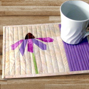 Könnte beinhalten: Ein weißer Stoffuntersetzer mit violetter Umrandung, auf dem eine violette und rosafarbene Blume mit braunem Zentrum gestickt ist. Der Untersetzer liegt auf einer Holzoberfläche mit einer weißen Tasse.