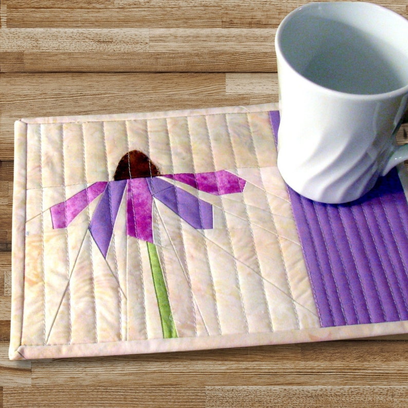 May include: A rectangular quilted fabric placemat featuring a pieced purple coneflower design with a brown center and green stem on a cream background, with a solid purple side panel and a white ceramic mug resting on the right side of the mat.