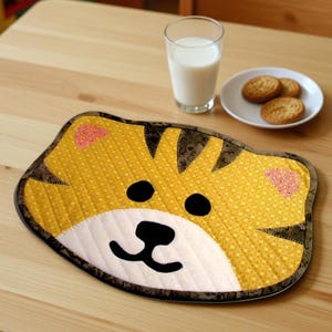 Pode incluir: Um lugar americano de tecido acolchoado em forma de rosto de tigre, com fundo amarelo estampado, listras pretas, orelhas cor-de-rosa e um focinho branco com nariz e boca pretos. O item possui uma borda marrom escura e detalhes costurados.