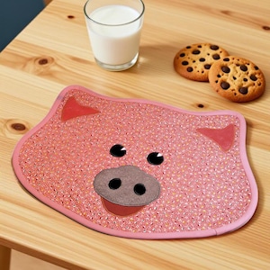 Op de afbeelding: Een roze placemat in de vorm van een varken met een klein bloemenpatroon, twee driehoekige oren, twee zwarte ronde ogen en een grote grijze snuit met twee zwarte neusgaten. De placemat heeft een gladde, afgeronde rand en een plat ontwerp.