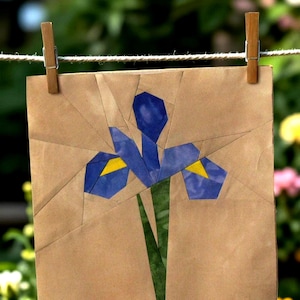 May include: A square paper collage featuring a geometric iris flower in shades of blue and yellow, set against a plain tan background. The artwork is suspended from a horizontal string by two wooden clothespins, showcasing sharp angular segments.