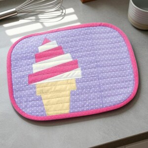 May include: A rectangular quilted potholder with rounded corners, featuring a lavender fabric background with a white polka dot pattern. The center displays a pieced fabric ice cream cone in shades of pink, white, and pale yellow.
