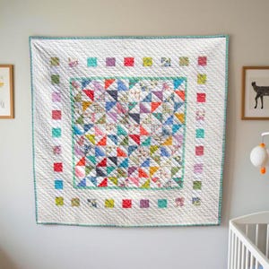 May include: A square quilt featuring a central panel of multi-colored triangle patches in various floral and geometric prints. The center is framed by a white border with diagonal quilting and a perimeter of small, colorful square fabric blocks.