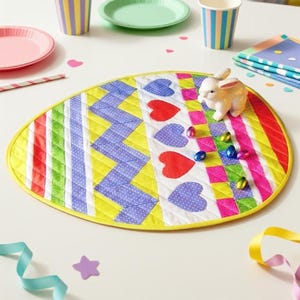 May include: An oval-shaped quilted fabric mat featuring a vibrant patchwork design of yellow, green, red, and purple stripes, chevron patterns, and heart shapes. A small white rabbit figurine and several colorful foil-wrapped chocolate eggs sit on top.