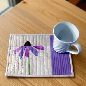 May include: A rectangular quilted fabric mat featuring a pieced purple and green coneflower design on a cream background with a solid purple side panel, paired with a white ceramic mug with a swirling textured surface and a curved handle.