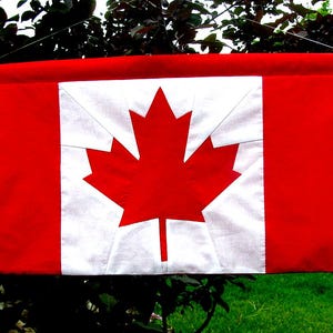 Pode incluir: Uma tapeçaria retangular de tecido com o design da bandeira canadense. A peça é construída com painéis de tecido vermelho e branco, exibindo um emblema central de folha de bordo vermelha sobre um quadrado branco, ladeado por duas faixas verticais vermelhas.