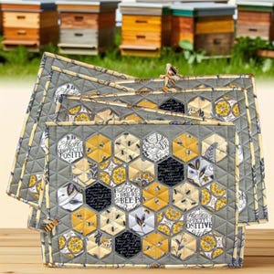 May include: A set of three quilted fabric mats featuring a hexagonal patchwork design in shades of yellow, grey, black, and white. The pattern includes bee illustrations, floral motifs, and text reading "Bee Respectful" and "Bee Positive" on a grey background.