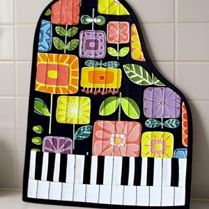 May include: A quilted oven mitt shaped like a grand piano, featuring a black background with a colorful pattern of stylized square flowers in shades of orange, yellow, pink, and purple. The bottom section displays a black and white piano keyboard.