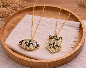 以下が含まれることがあります： 2つの金色のネックレス。ペンダントは、フットボール型と「SAINTS」の文字が入った盾型。どちらのペンダントも黒いフルール・ド・リスのデザインで、小さな透明な石がちりばめられています。