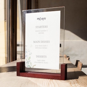 Könnte beinhalten: Ein durchsichtiges Acryl-Menüschild mit einem Sockel aus dunklem Holz. Die weiße Papiereinlage zeigt den Text menu, STARTERS, MAIN DISHES und DESERTS sowie grüne Aquarell-Blattillustrationen auf der linken Seite des Schildes.