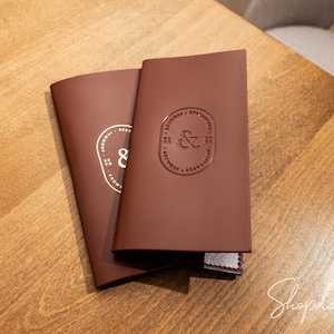 May include: Two rectangular dark brown leather menu covers featuring a debossed oval emblem with an ampersand symbol in the center. The covers are smooth with clean edges and a small white fabric tag visible at the bottom corner of the top cover.
