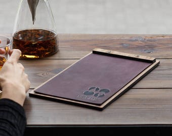Puede incluir: Un portamenús de cuero morado oscuro con una barra superior de madera y dos tornillos metálicos, que presenta un logotipo grabado en la parte inferior izquierda. Junto a él hay una jarra de cristal transparente con líquido ámbar y un vaso.