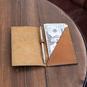 May include: Tan leather wallet with a pen and cash on a wooden table. The wallet has a triangular pocket holding several twenty-dollar bills. A silver pen is secured with a leather loop. The wallet is open, revealing the interior.