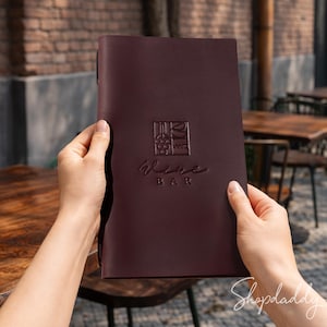 以下が含まれることがあります： 幾何学模様の正方形のロゴと「Wine Bar」の文字が型押しされた、ダークバーガンディ色のレザー製メニューカバー。長方形のカバーは滑らかでマットな質感で、正面がはっきりと見えるように垂直に保持されています。