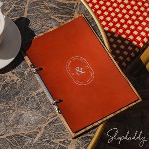 Puede incluir: Un menú rojo con marco de madera y anillas plateadas descansa sobre una mesa de mármol junto a una taza de café blanca. El menú tiene un logotipo blanco con el símbolo de la &.